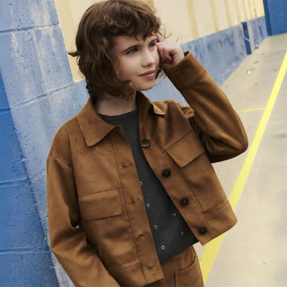 A girl model wearing a brown suede-like jacket over a dark gray top, standing outdoors against a blue and yellow wall.