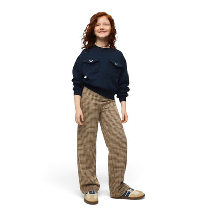 A young woman with red hair stands against a solid white background, wearing a navy blue sweatshirt with chest pockets, brown and beige plaid trousers, and beige slip-on shoes.
